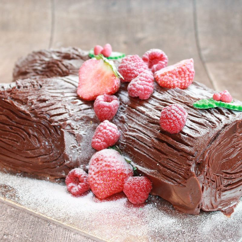 A festive yule log cake decorated with strawberries and raspberries on top, frosted with white icing and sprinkled with powdered sugar.