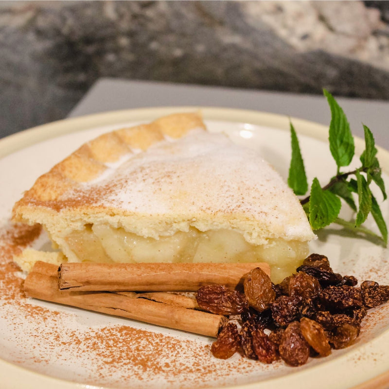 A homemade American apple pie topped with a layer of dusted cinnamon, accompanied by cinnamon sticks and raisins on a white plate.